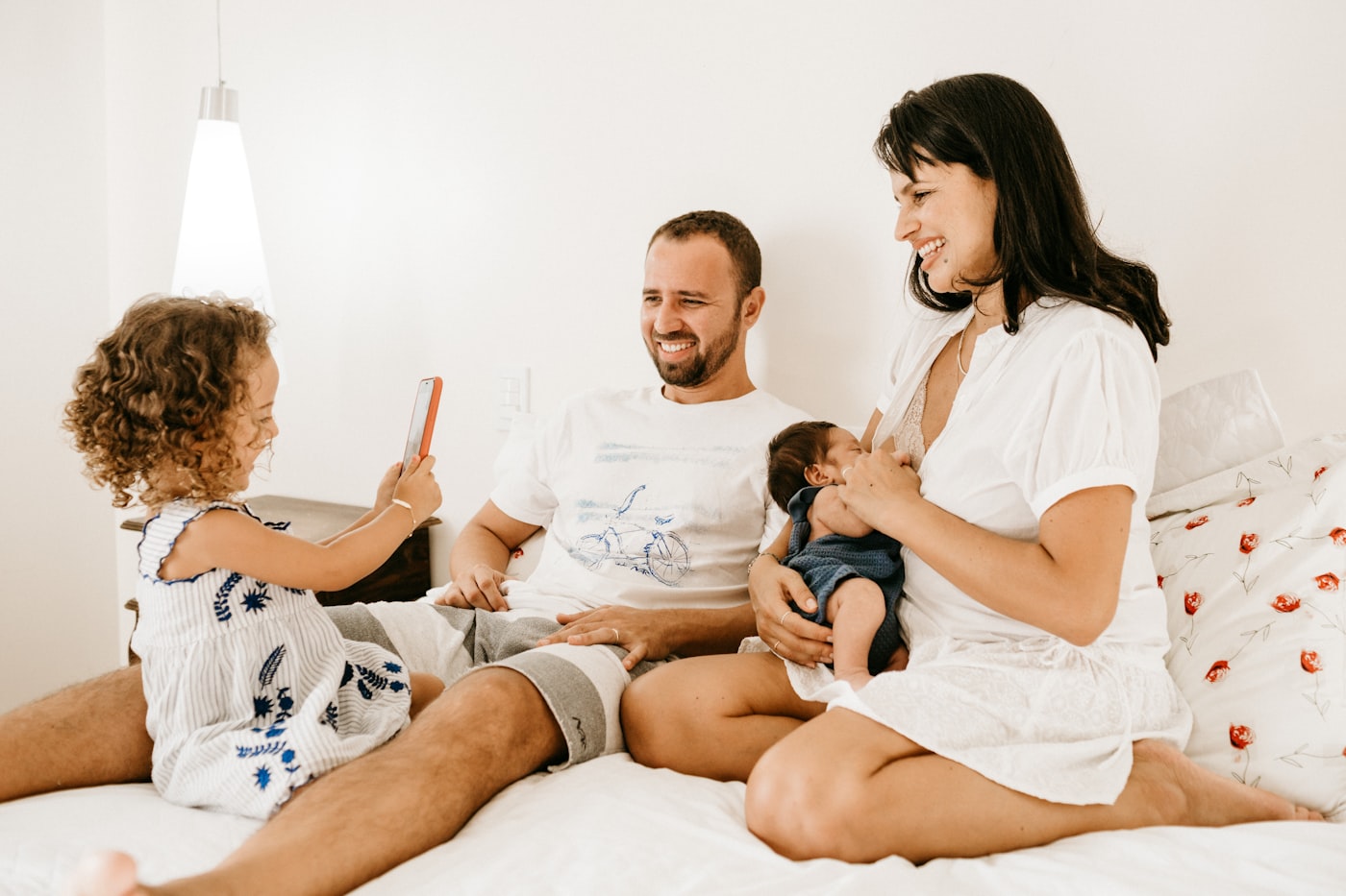 Two parents sitting close on a bed with their baby, laughing in soft morning light.