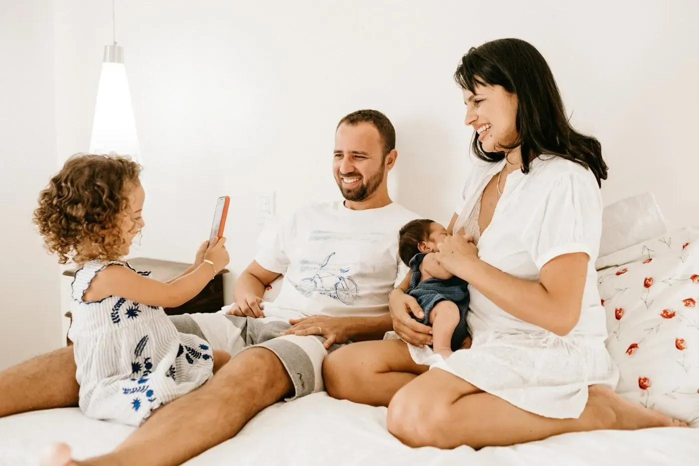 Two parents sitting close on a bed with their baby, laughing in soft morning light.